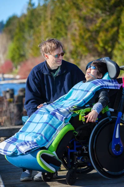 Niepełnosprawnych chłopak na wózku inwalidzkim, rozmawia z ojcem w parku nad jeziorem — Zdjęcie stockowe