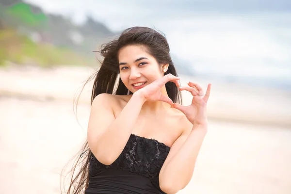 Adolescente chica de pie en la playa sonriendo, las manos formando forma de corazón —  Fotos de Stock