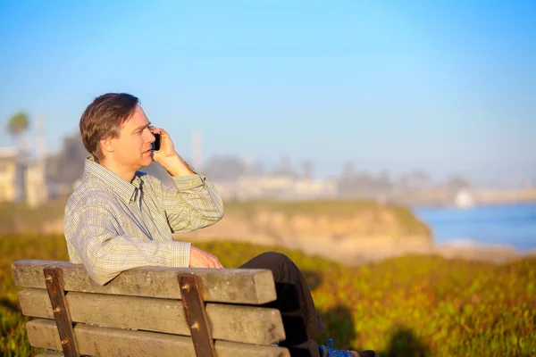 Businessman talking on cellphone outdoors,  park bench by ocean — Stock Photo, Image