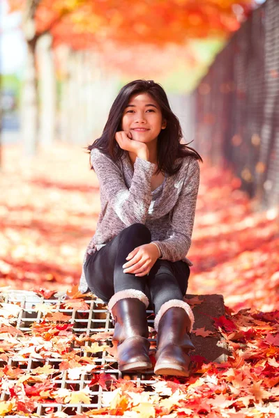 Sonbaharda renkli akçaağaç altında oturan biracial genç kız — Stok fotoğraf