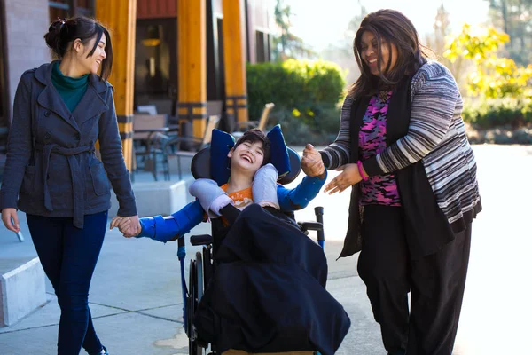 Мальчик-инвалид в инвалидной коляске держит за руки санитаров на прогулке — стоковое фото