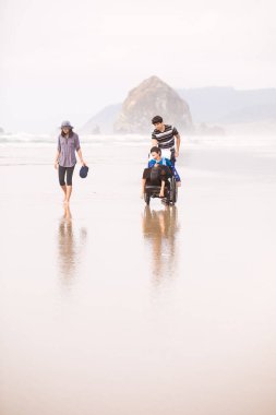 Little disabled boy in wheelchair with family walking along beac