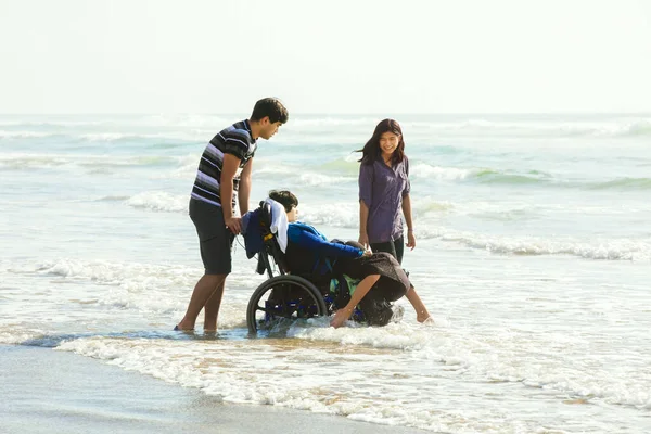Pequeño niño discapacitado en silla de ruedas con familia caminando a lo largo de la playa —  Fotos de Stock
