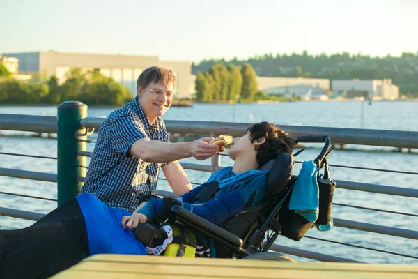 Padre alimentación hijo discapacitado en silla de ruedas hamburguesa por urbano la —  Fotos de Stock