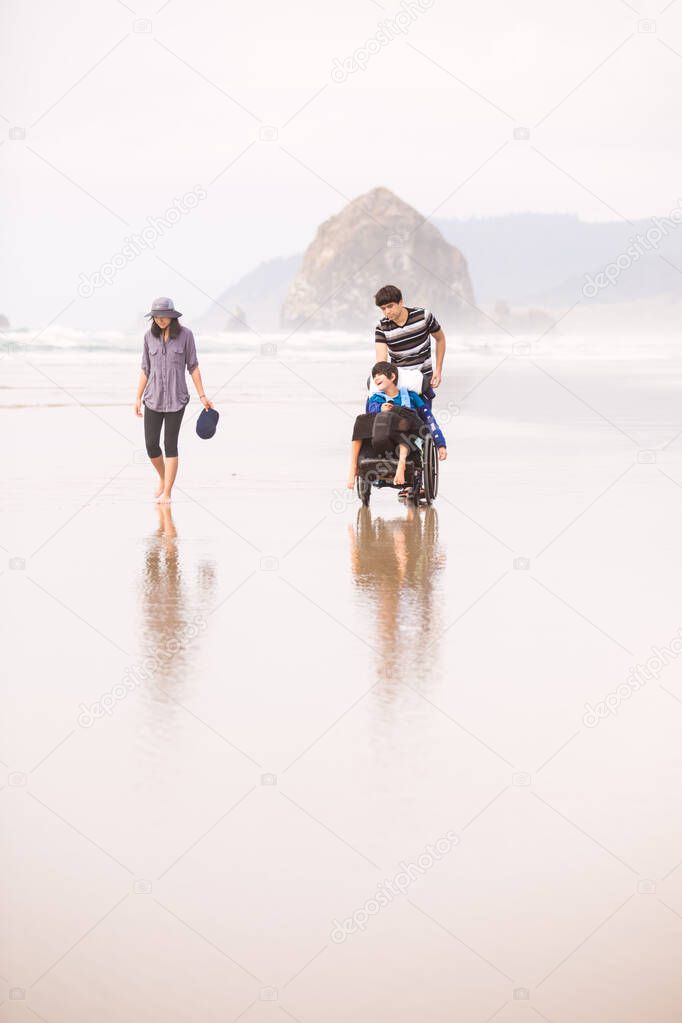 Little disabled boy in wheelchair with family walking along beac