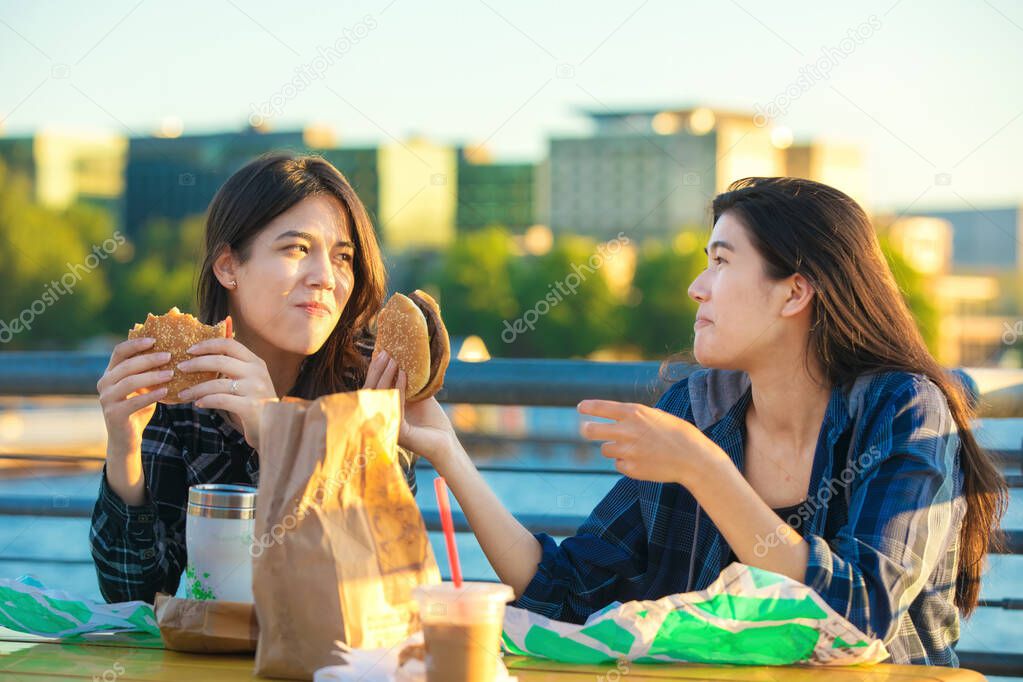 Two teen girls eating hamburgers outdoors by lake, urban backgro