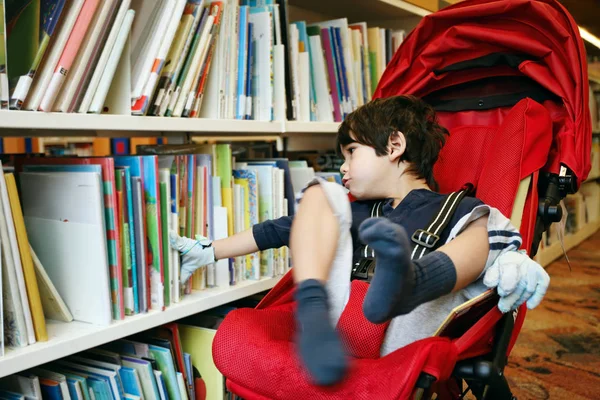 Niepełnosprawny chłopiec w czerwonym wózku wybierający książki w bibliotece — Zdjęcie stockowe