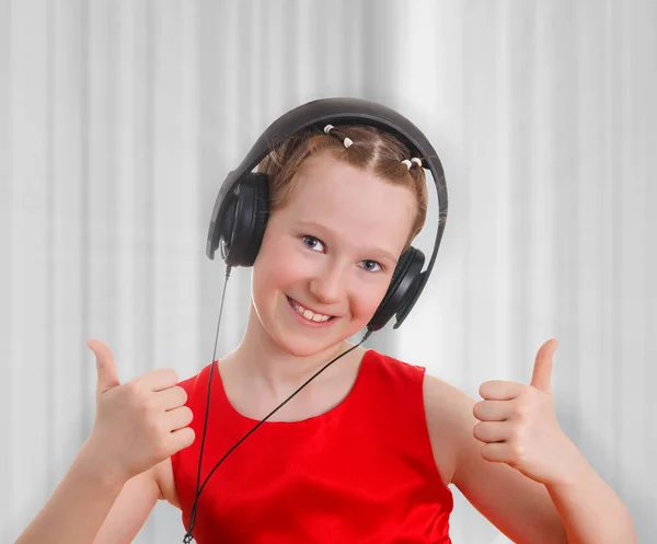 Linda chica adolescente en un vestido rojo escuchando música en los auriculares y mostrando un gesto hacia arriba pulgares, como . — Foto de Stock