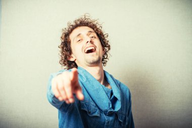 Kıvırcık genç adam işaret parmağı göstererek gülüyor. Gri bir arkaplanda