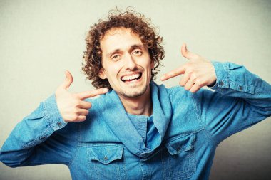 Kıvırcık genç adam dişçiden sonra işaret parmağını göstererek gülüyor. Gri bir arkaplanda