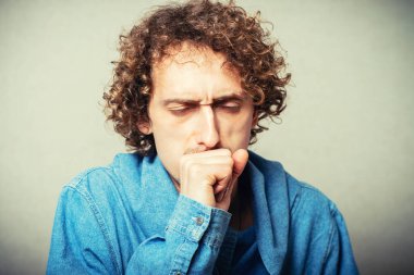 Kıvırcık genç adam eli ağzında öksürüyor. Gri bir arkaplanda