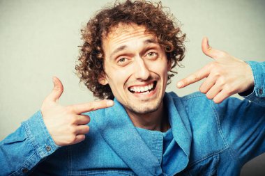 Kıvırcık genç adam dişçiden sonra işaret parmağını göstererek gülüyor. Gri bir arkaplanda