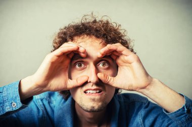 Kıvırcık genç adam gözlerinin etrafındaki dürbünü elleriyle yapıyor. Gri bir arkaplanda