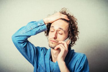 Kıvırcık genç adam telefonda konuşuyor, kötü haberler alıyor. Gri bir arkaplanda