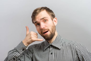Gri arka planda çizgili gömlekli genç bir adam. Elleriyle bir telefon hareketi yapıyor.