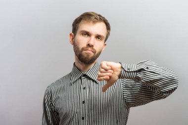 Gömlekli ve kravatlı genç bir adamın portresi kameraya bakıyor ve gri arka planda dikilirken baş parmaklarını aşağı gösteriyor.