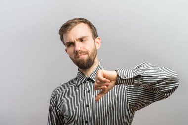 Gömlekli ve kravatlı genç bir adamın portresi kameraya bakıyor ve gri arka planda dikilirken baş parmaklarını aşağı gösteriyor.
