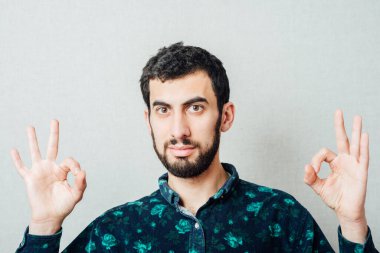 Her şey yolunda! Tişörtlü mutlu genç adam Gri arka planda dururken işaret veriyor ve gülümsüyor