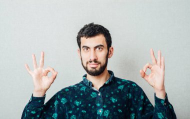 Her şey yolunda! Tişörtlü mutlu genç adam Gri arka planda dururken işaret veriyor ve gülümsüyor