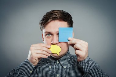 Gri arka planda ağzını ve gözlerini kapayan bir kağıt parçasıyla genç bir adamın yakın plan portresi.