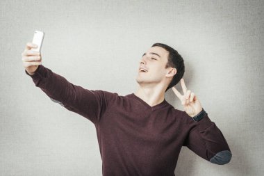 Yakışıklı, genç bir adam elinde kamerayla özçekim yapıyor ve gri arka planda dikilirken gülümsüyor.