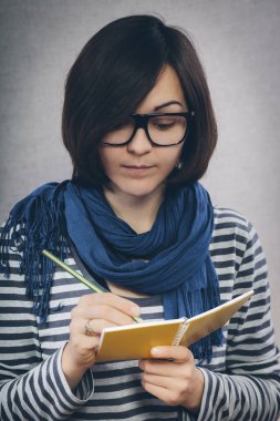 Gözlüklü genç kadın deftere bir şeyler yazıyor.