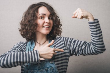 Kadın şakacı bir şekilde elindeki kasları gösteriyor. Gri bir arkaplanda.