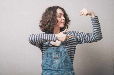 Kadın şakacı bir şekilde elindeki kasları gösteriyor. Gri bir arkaplanda.