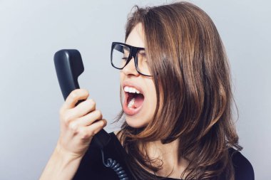 Ofisi ya da ev telefonu olan kadın küfrediyor, çığlık atıyor. Gri bir arkaplanda.