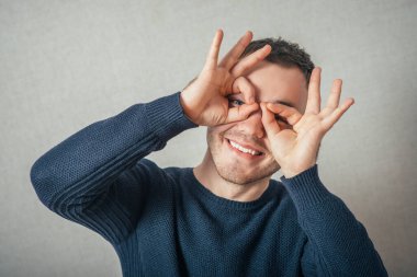 Bir adam parmaklarının gözlüklerinden bakar. Gri bir arkaplanda.