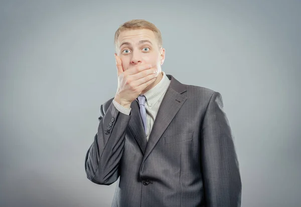 Portrait of afraid businessman wearing suit covering his mouth with his hand over grey background.