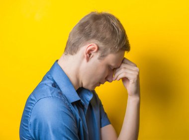 Sarı arka planda mavi gömlekli genç adam. Başının altında iki el yorgunluk, öfke, yanlış anlaşılma gösteriyor. Jest. fotoğraf çekimi.