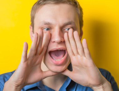 İki eliyle ağzını kapatıyor ve yüksek sesle bağırıyor. Sarı arka plandaki genç adam. Fotoğraf. Erkekler yüksek sesle birilerini arıyor..