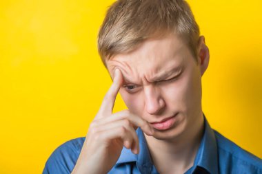 Young man close-up in a blue shirt on a yellow background, close one eye, pissed off. Mimicry. Zhest.Foto
