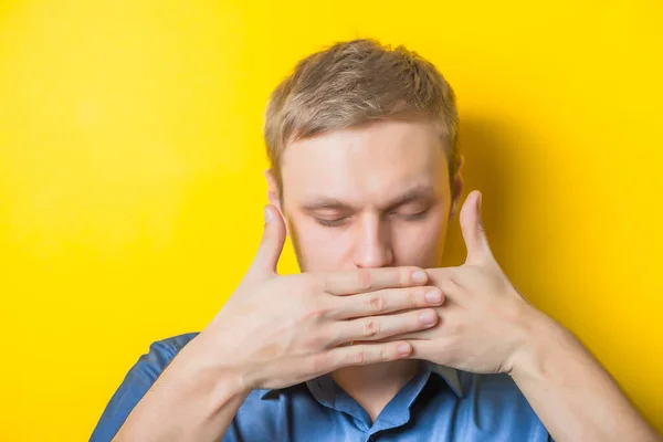 Blond young man closed his mouth with his hands, does not speak. Silence Gesture. Close portrait. Isolated yellow background. photo
