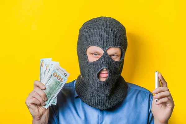 男はマスクの泥棒と黄色い背景の青いシャツを閉じてカメラをじっと見てお金と電話を持ってた ミミイキー ジェスチャー 写真撮影 — ストック写真