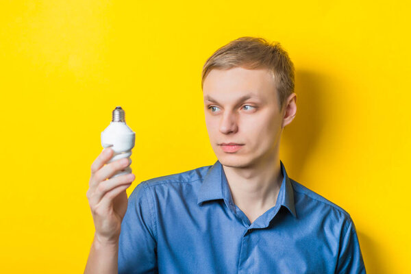 Young man close-up in a blue shirt on a yellow background holding a light bulb energy saving, looking at her. Mimicry. Gesture. photo