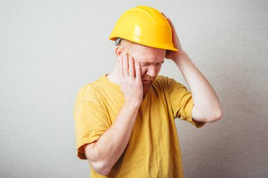 Sarı tişörtlü, sarı miğferli, sakallı, gri arka planlı bir adam.