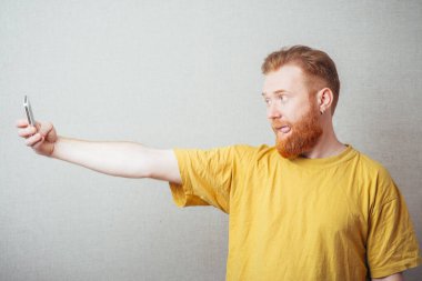 Sakallı, sarı tişörtlü, gri arka planlı bir adamda cep telefonunda senin fotoğrafın var.