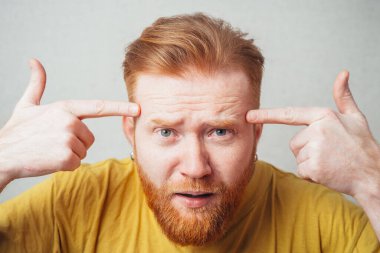 Sarı tişörtlü, sakallı, gri arka planlı bir adam öfkeden kuduruyor ve kafasında işaret parmağı gösteriyor.