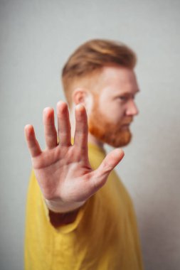 Sarı tişörtlü, sakallı, gri arka planlı bir adam öfkeden kuduruyor ve durduğunu gösteriyor.