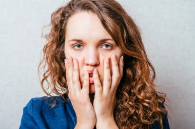 Kıvırcık kız şaşırdı, yüzünün yanında korkmuş eller vardı. Gri arkaplan.