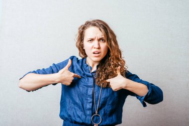 Kıvırcık kız göğsündeki işaret parmağını gösteriyor. Gri arkaplan.