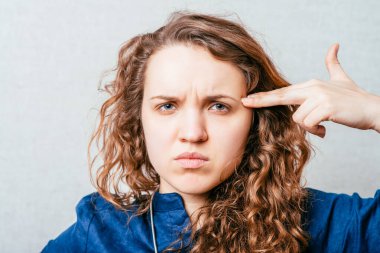 Kıvırcık kız sinirli intiharı, silah şeklindeki bir tapınağa parmağını simüle eder. Gri arkaplan.