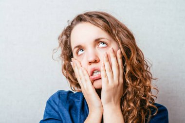 Kıvırcık kız sinirli, elleri yanaklarında. Gri arkaplan.