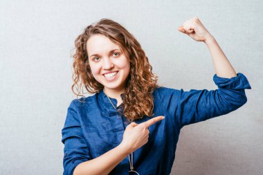 Kıvırcık kız şakacı bir şekilde kolunun gücünü gösteriyor. Gri arkaplan.