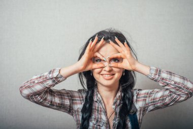 Parmaklarının çerçevesinden bakan, gözlük yapan, parmaklarının dürbünü olan bir kadın. Gri bir arkaplanda.