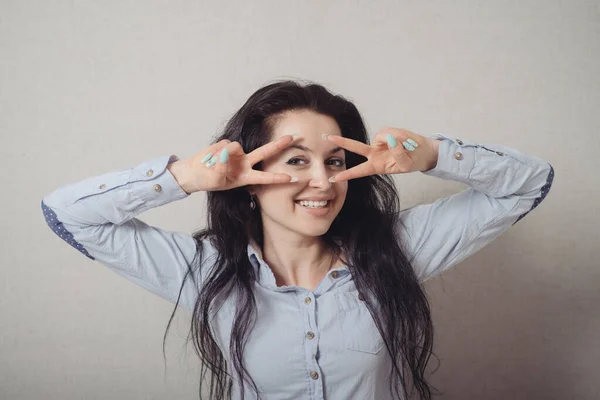 一个女人通过他的手指看 一个胜利的姿态 灰色背景 — 图库照片