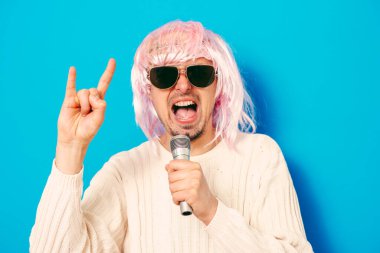 Peruklu ve güneş gözlüklü bir adam mikrofona şarkı söylüyor..