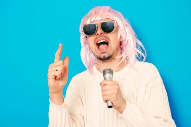Peruklu ve güneş gözlüklü bir adam mikrofona şarkı söylüyor..
